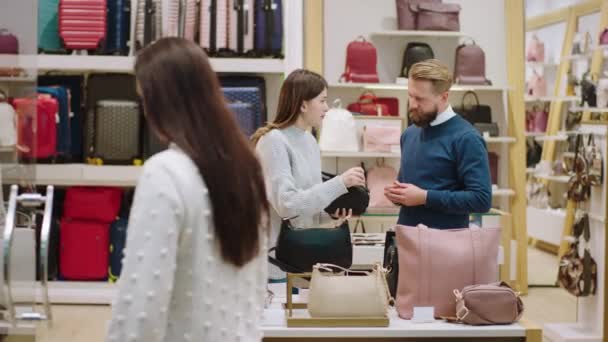 У сучасному магазині аксесуарів продавець розмовляє з красивою клієнткою молода жінка, яка тримає сумку і обговорює зосереджену з чоловіком — стокове відео