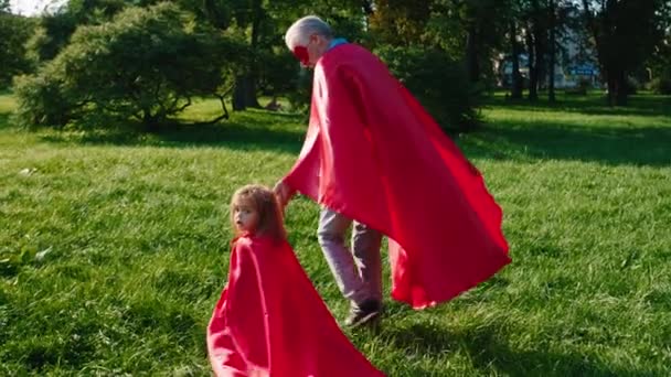 公園のカメラの前で草を一緒に歩く祖父と彼の小さな美しい姪彼らは素晴らしいスーパーヒーローのスーツを着て — ストック動画