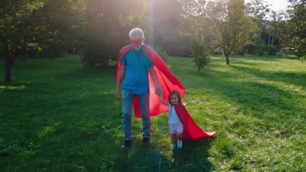 No meio do parque carismático homem velho olhando e sua filha pequena bonito andando um em um terno de super-herói juntos de mãos dadas — Vídeo de Stock