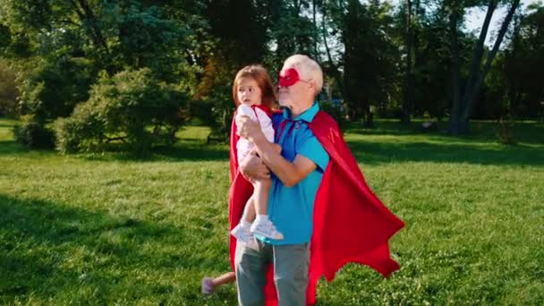 公園の真ん中で飛行機で遊んでいる間、カメラの祖父と彼の姪は甥と一緒にスーパーヒーローのスーツを着て — ストック動画