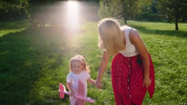 In the sunny day in the park happy young mommy with her daughter running in front of the camera with a pink airplane — Stockvideo