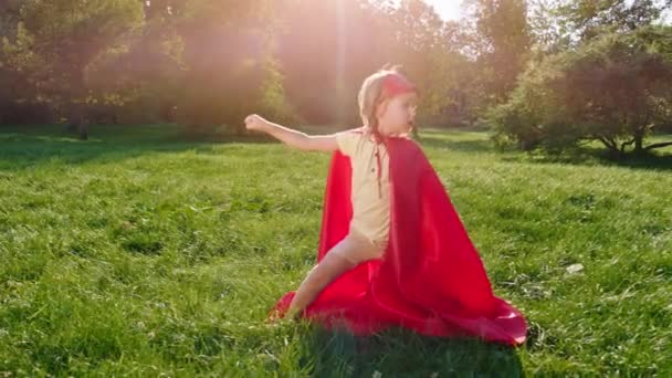 Ελκυστικό και αστείο χαριτωμένο αγοράκι με κόκκινη στολή υπερήρωα που ποζάρει μπροστά στην κάμερα στη μέση του πάρκου — Αρχείο Βίντεο