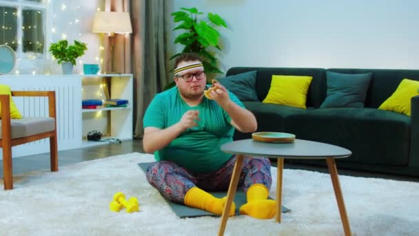 家で働く肥満の男のためにクランチをしながら食べるのを停止します。半分のハンバーガーを食べながらリビングルームの床に座って — ストック動画