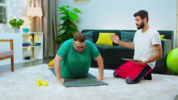 거실에 있는 두 명의 좋은 친구들 이 일 을 시작 했습니다. 뚱뚱 한 남자가 무게를 줄이기 위해 운동을 하는 동안 그 의 친구는 디지털 태블릿을 사용하여 — 비디오