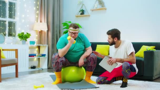 En el chico de la sala de estar ayudando a su amigo obeso a hacer los ejercicios correctos para perder el peso que utiliza la tableta digital para monitorear el pulso — Vídeo de stock