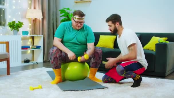 Genç adam, şişman arkadaşının evde salonda vücut çalışmasına yardım ediyor. — Stok video