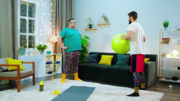 In the living room funny fat guy doing workout with his partner he pushing the sport ball with his big belly — стоковое видео
