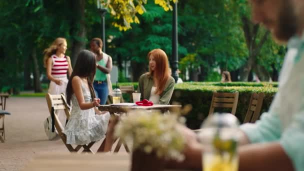 In the middle of the park at the cafe beautiful two ladies have a conversation while other ladies black woman and blonde hair lady came to their friends they hugging each other and enjoy the time — Vídeo de Stock