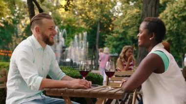 A beautiful black so man with short hair is drinking wine with a handsome man who has a beard, they are in a park with a beautiful fountain. Shot on ARRI Alexa Mini.
