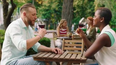 a couple is at the park sitting down at a table, sharing a glass of wine and cheering, they taste the wine and don t like it as they laugh it off