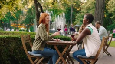 An angelic looking woman with ginger long hair is sharing and cheering some wine with her African friend, they are sitting at a table and surround by the park