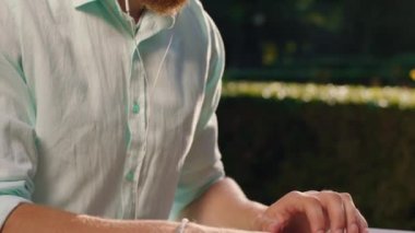 Kulaklığı takıp bilgisayarda çalışırken gülümseyen yakışıklı bir adamın yakın plan fotoğrafı. Arka planda ağaçlar var ve güneş onun üzerinde parlıyor.