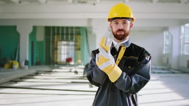 Buen tipo trabajador de la construcción con sus guantes protectores delante de la cámara y posando con las manos cruzadas que de pie en un gran edificio de espacio en la construcción — Vídeo de stock