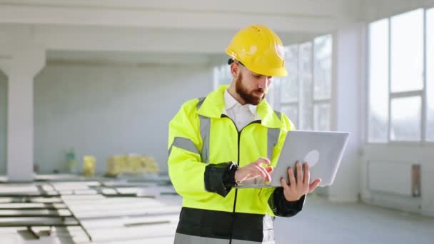 Charismatický inženýr na staveništi ve speciální uniformě držící notebook a pracující soustředěný na stavební plán — Stock video