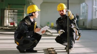 In the large construction site the man constructor worker and the woman engineer with digital tablet analysing the plan of work they looking at the heavy building materials
