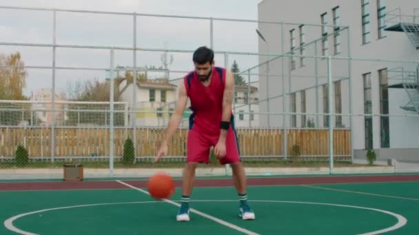 Gran cancha de baloncesto frente a la cámara hombre en traje deportivo rojo jugando concentrado con una pelota — Vídeos de Stock