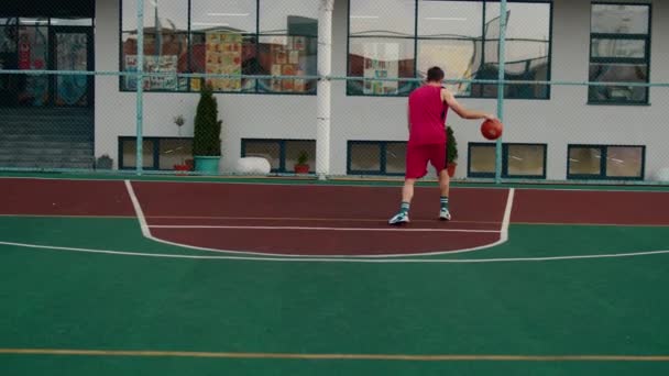 Красивий хлопець у червоному спортивному костюмі грає в баскетбол на стадіоні він кидає м'яч у кошик — стокове відео