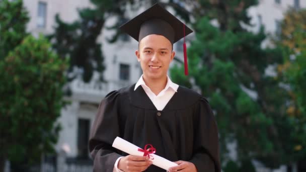 Smiling large happy guy graduate and holding his diploma in front of the camera he wearing graduation suit and cap — Stock video