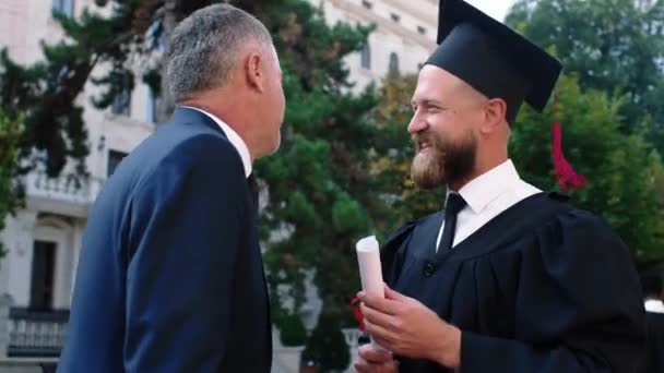 Closeup to the camera charismatic graduate man have a conversation with his dad very excited and happy on his graduation day — стоковое видео