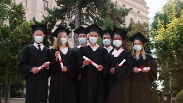 Charismatic multiracial graduates students outside of the college posing in front of the camera in the pandemic of coronavirus they holding diplomas in hand and wearing protective mask — стоковое видео