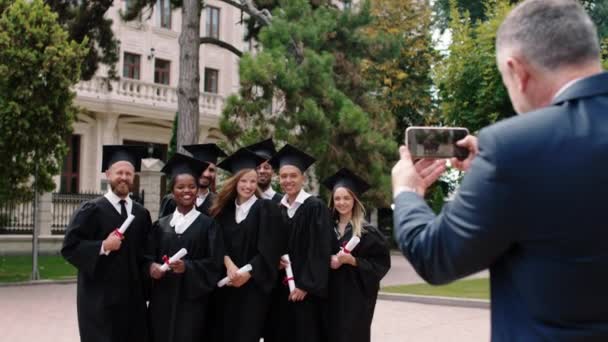 Diplomaosztó nap egy csoport multiracionális végzős diákok pózol egy nagy mosollyal nagyon izgatott előtt az okostelefon a főiskolai parkban az egyik szülő öregember fényképez — Stock videók