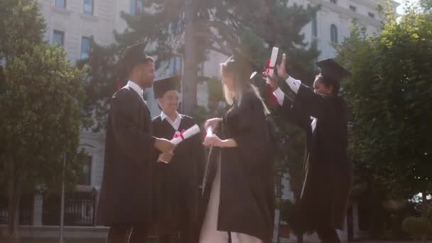 Excited and happy students graduates after they take their diplomas dancing and jumping in the college garden wearing graduation suits and caps — Stockvideo