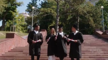 Very excited group of multiracial graduates students walking down the stairs outside of the college building they are happy holding diplomas and wearing graduation caps