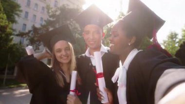 In the college garden multiracial graduates students young women and one guy using the camera to make a selfie video they are smiling large looking straight to the camera posing and holding diplomas