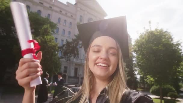 Excited great looking lady graduate she using the camera to make a selfie video she speaks something on the camera and showing her diploma in the college garden — Vídeos de Stock