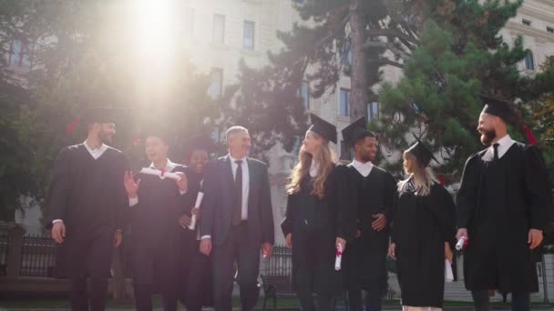 В саду колледжа группа многорасовых студентов заканчивает выпуск перед камерой, гуляя вместе с директором колледжа и улыбаясь с большим волнением — стоковое видео