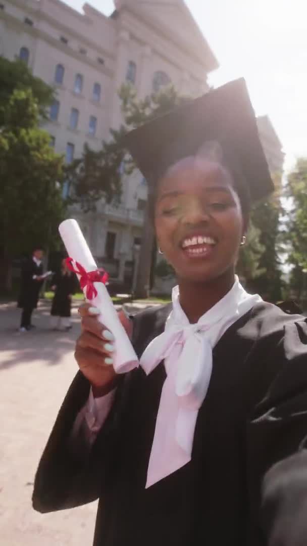 Beautiful black woman student graduate using the camera to make a selfie video for her family she speaks something and holding in hands her diploma after the graduation — kuvapankkivideo