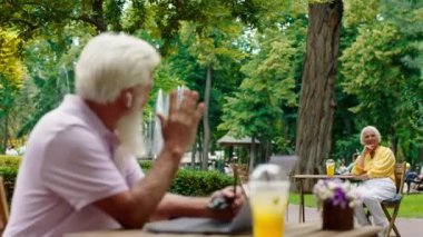 In the middle of the park at the cafe old man with a white chin he say hi to a beautiful woman who sitting beside him at the cafe as well