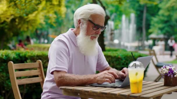 Charismatic old man with sunglasses working at the cafe typing something on the laptop in the middle of park and listen musing from earphones phones — Stockvideo