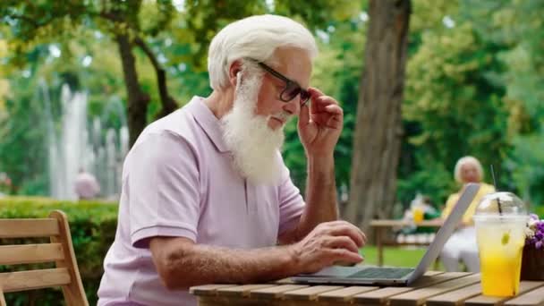En un día de verano, un viejo atractivo con una gran barba blanca trabajando duro en su computadora portátil se quita las gafas de sol, tiene una cara cansada. — Vídeos de Stock