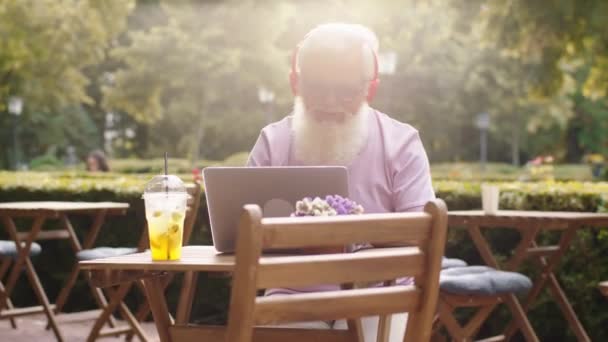 Happy and excited old man good looking typing something on the laptop and listen music from headphones and enjoy the sun in summer day — Stock video