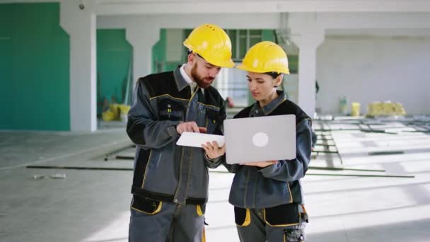 Charismatic woman engineer inside of construction building analysing together with her assistant the plan of work using the laptop and digital tablet they wearing safety helmets and special uniforms — Vídeo de Stock