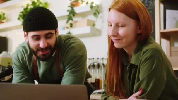 Sıcak kafeteryada harika bayan barista ve yakışıklı barista laptopun üzerinde çalışırken sohbet ediyorlar. Büyük bir gülümseme konseptiyle iyi bir takım oluşturuyorlar. — Stok video