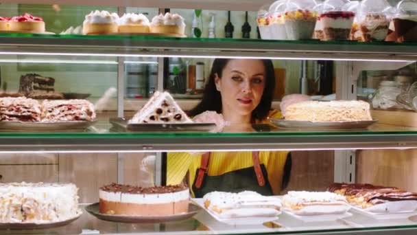 Schöne kaukasische Frau mit einem hübschen Lächeln die Besitzerin des Cafés, das in ihrem Café arbeitet, arrangierte sie sorgfältig die Desserts aus dem Vitrinenschrank — Stockvideo