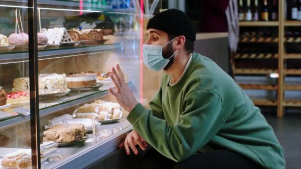 Chico guapo con máscara protectora cliente mirando la nevera escaparate en el café de la panadería para elegir el postre favorito en la pandemia de coronavirus — Vídeos de Stock