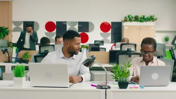 Dos trabajadores africanos deslumbrantemente atractivos están en una gran oficina iluminada, que están ayudando a los demás con el trabajo y hablando mientras están en sus mesas — Vídeos de Stock