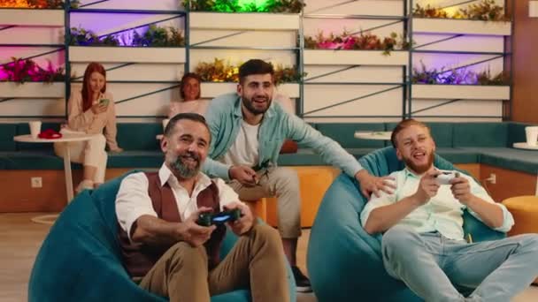 Twee jongere mannen en een ouder uitziende man spelen videospelletjes terwijl ze in zitzakken zitten en elkaar aanmoedigen. Arri Alexa Mini. — Stockvideo
