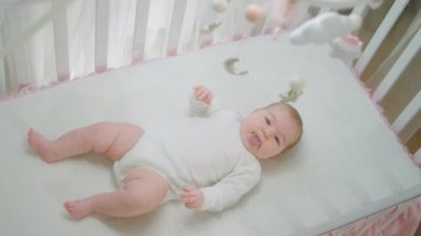Bebek karyolasında tek başına, elleri ve bacaklarıyla oynayıp bebek ve çocukluk bebek oyuncakları konseptini araştıran inanılmaz tatlı bir kız.
