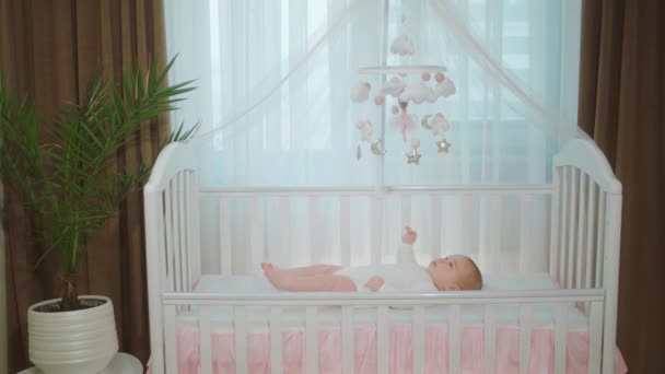 아주 예쁜 여아가 카메라 앞에 혼자 누워 아기 침대에 누워서 아기 장난감을 가지고 놀고 있는 모습 — 비디오