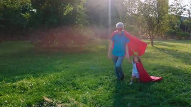Yakışıklı yaşlı adam ve küçük kızı kırmızı bir süper kahraman kıyafeti giyip parkın ortasında çimlerin üzerinde yürüyorlar. — Stok video