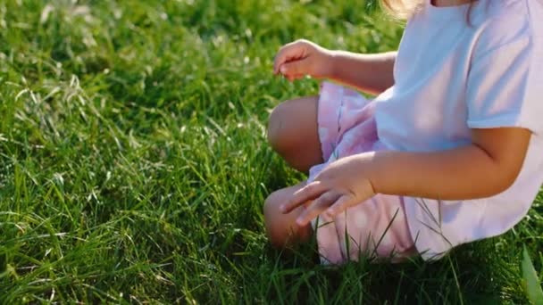 Dole na trávě roztomilá holčička hraje s trávou a dívá se přímo na kameru směšně se usmívá a těší čas v parku — Stock video
