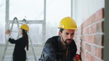 Güvenlik kasklı, dijital tabletli kadın kameranın önünde karizmatik işçi duvarları incelerken bazı inşaat resimlerini çekmek için bekliyor.