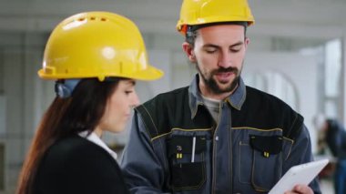 İnşaat alanındaki kameraların önünde çekici erkek müteahhit ve inşaat planını analiz eden denetçi kadın güvenlik kaskı ve üniforma giyiyorlar.