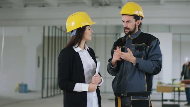 Em um grande local de construção bela mulher engenheiro eo trabalhador construtor homem com capacetes de segurança discutindo sobre o plano de construção juntos — Vídeo de Stock