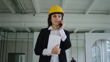 İnşaat alanında kameraman kadın mühendisin önünde yürüyor ve elinde dijital tabletle güvenlik kaskı takarak akıllı telefonuyla konuşuyor.