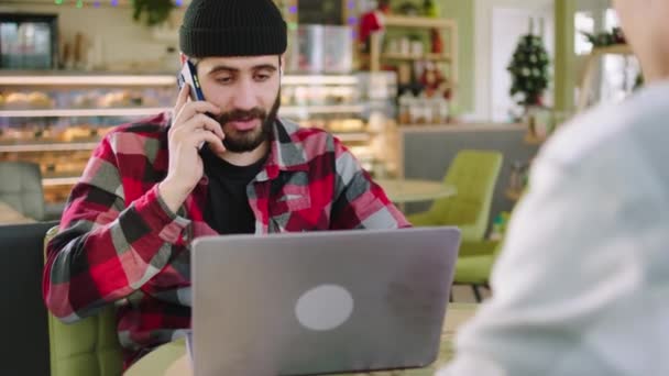 Vor der Kamera sitzt der attraktive Kerl mit Laptop konzentriert mit seinem Freund in einem modernen Coffeeshop und antwortet mit seinem Smartphone. 4k — Stockvideo